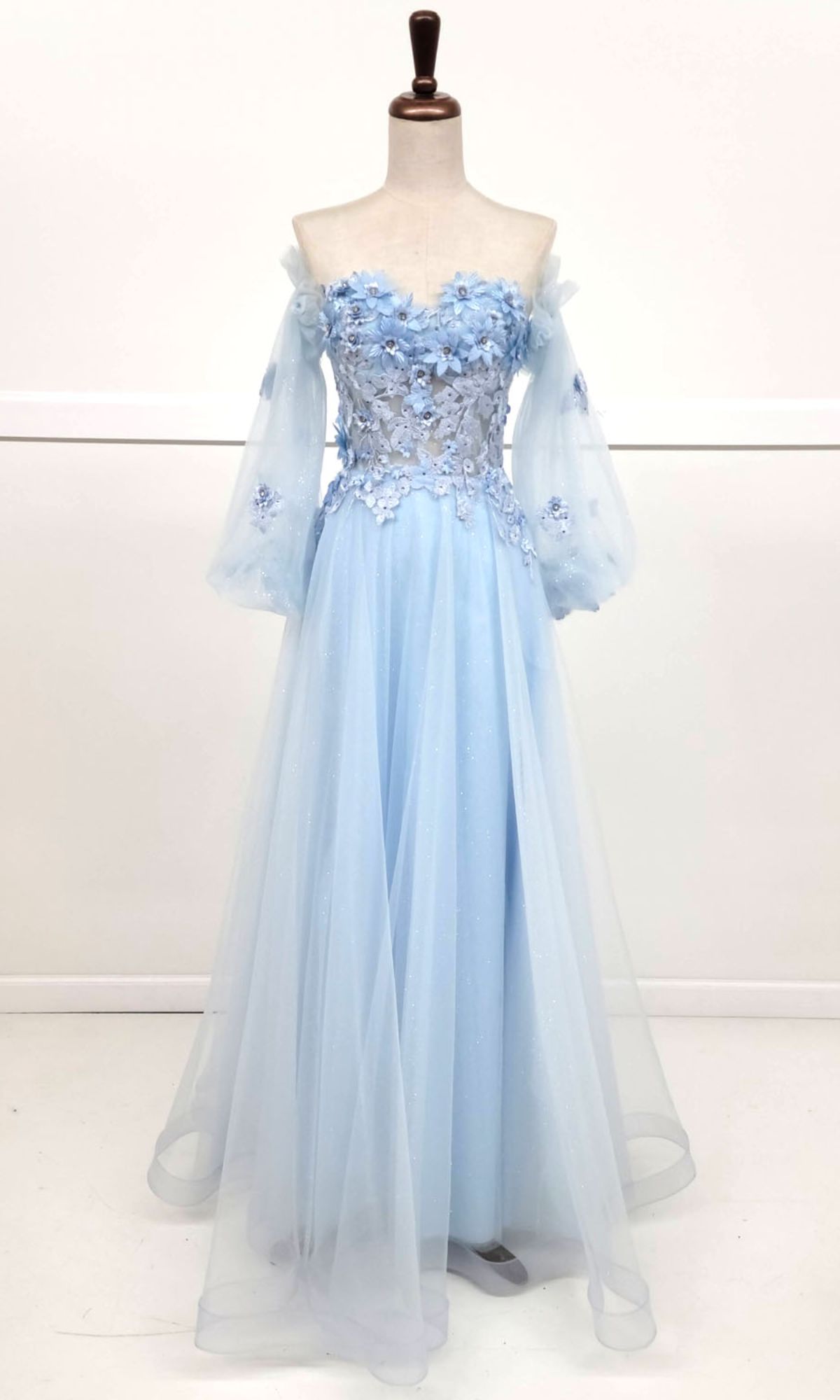 The Chicas CHF3096 Long Sleeve Long Prom Ball Gown features a light blue off-the-shoulder design with a floral appliqué bodice, sheer puff sleeves, and a flowing tulle skirt, showcased on a dress form against a white background.